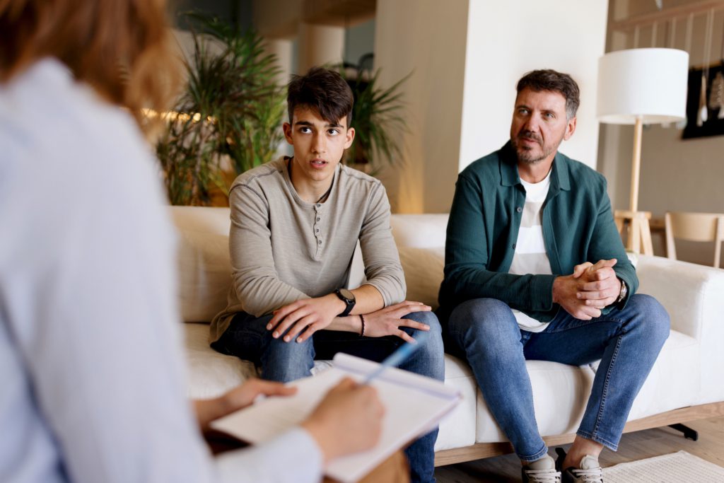 A teenage boy and his father sit on a sofa while listening to a partially unseen female therapist.
